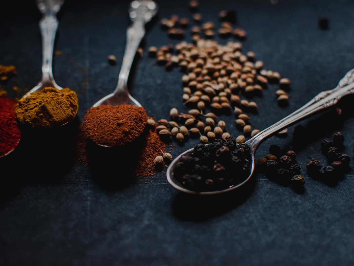 Ground and whole spices arranged in metal spoons on a dark surface